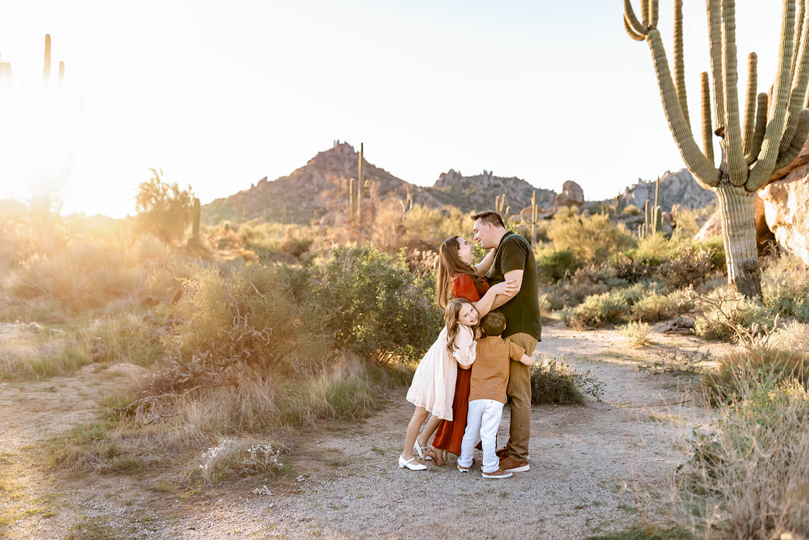 Mom and dad kiss while kids hug them in a desert family photo session in beautiful complementary outfits