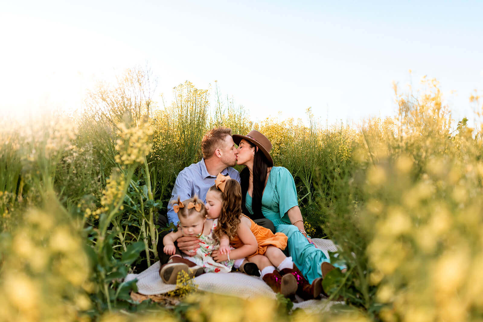 Family of four sit in a field of yellow flowers with mom and dad kissing while little girls snuggle in front of them.