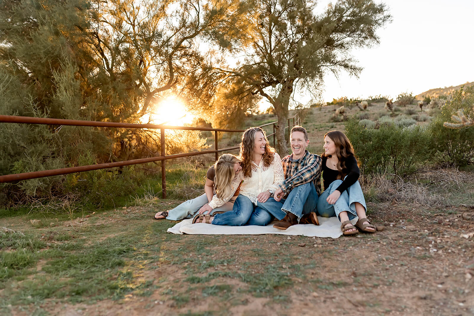 Family of four sit on a beige blanket with a golden sunset and trees behind them in Phoenix