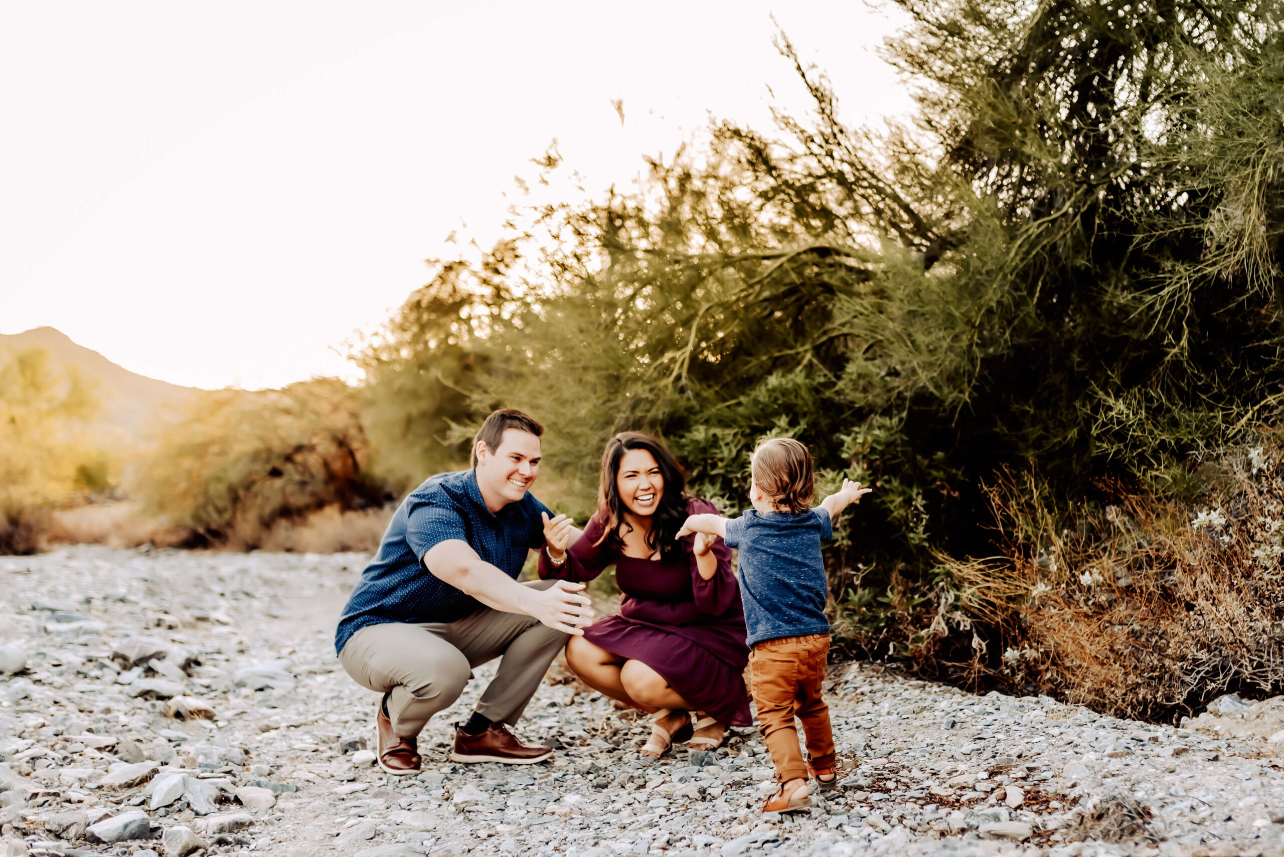 Mom and dad crouch down on gravel path waiting to embrace their toddler who runs towards them