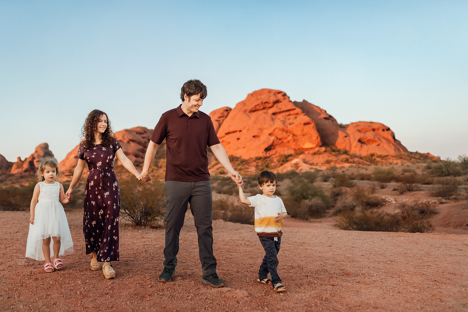 Family of four hold hands and walk with buttes glowing in the sunset at Papago Park, Phoenix