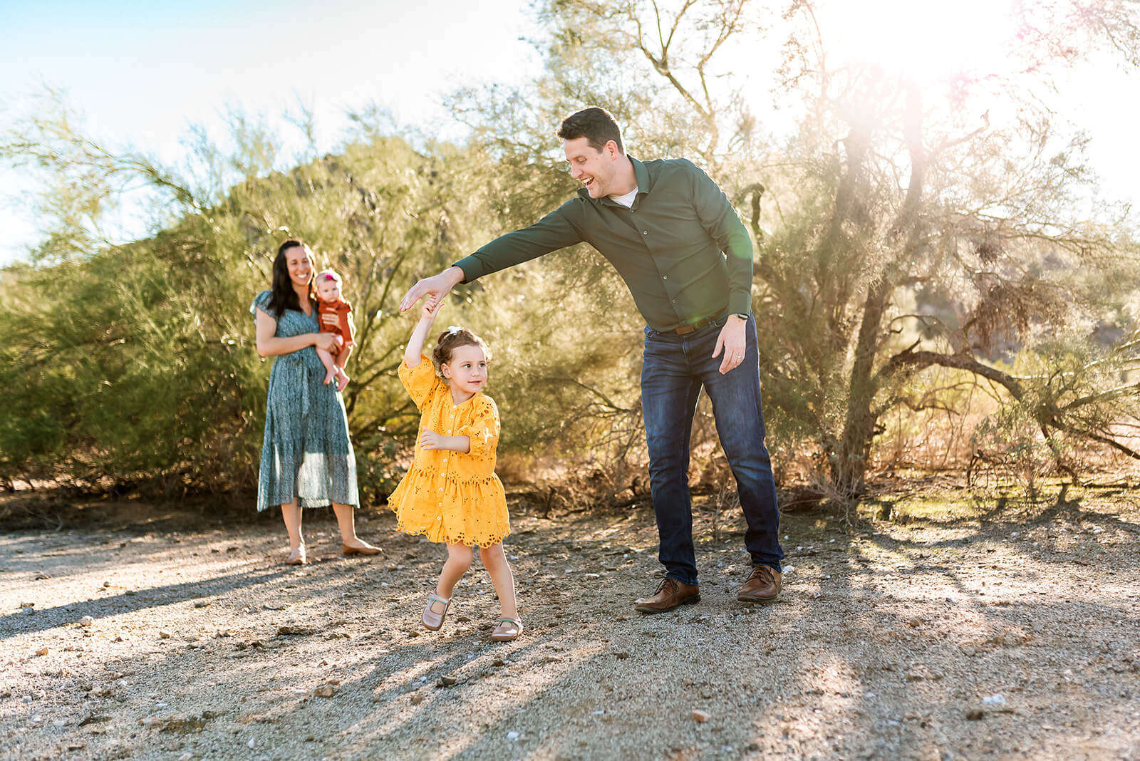 Dad twirls little girl wearing yellow while mom holds baby in the background in a desert photo session in Scottsdale.