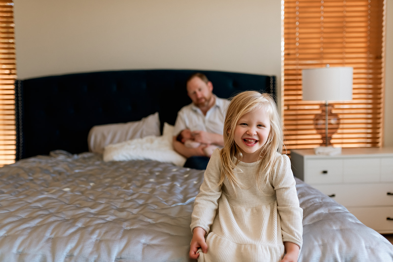 Little girl sits on the edge of the bed smiling gleefully as her dad holds her newborn baby sister behind her.