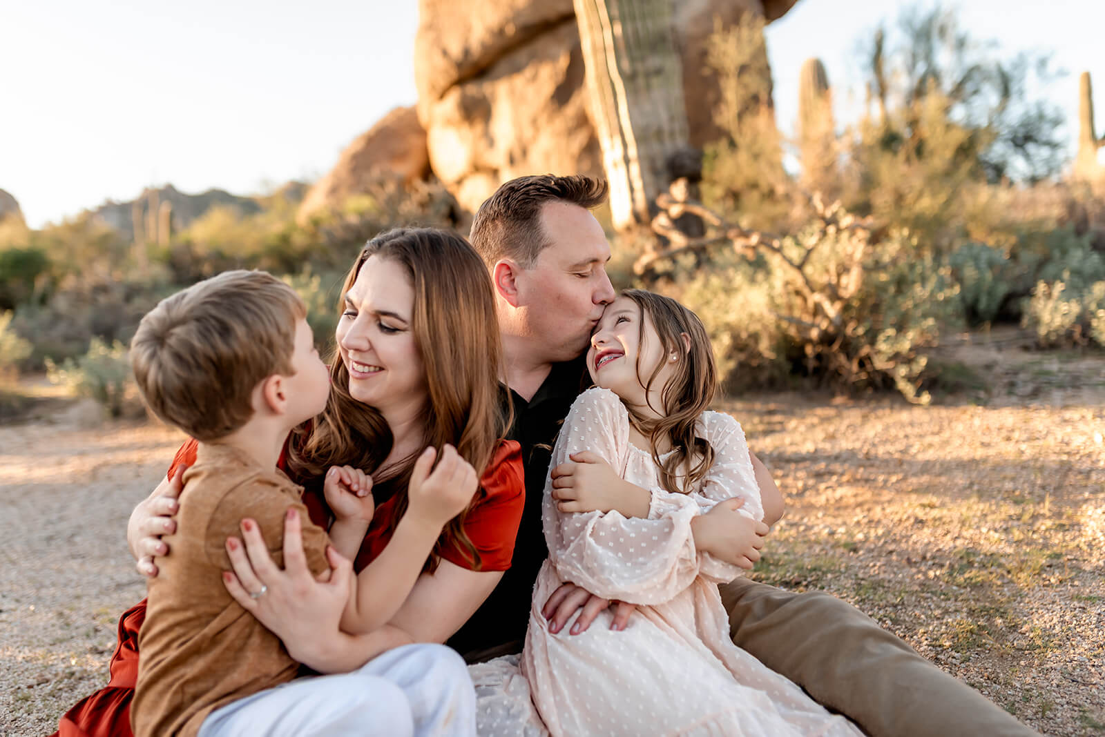 Family of four look at each other and laugh while sitting on a blanket in the Scottsdale desert.