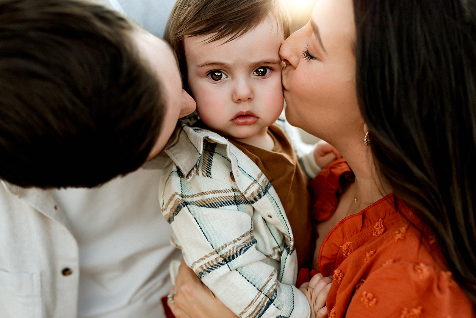 Little boy stares intently at the camera while mom and dad give him kisses on his cheeks.