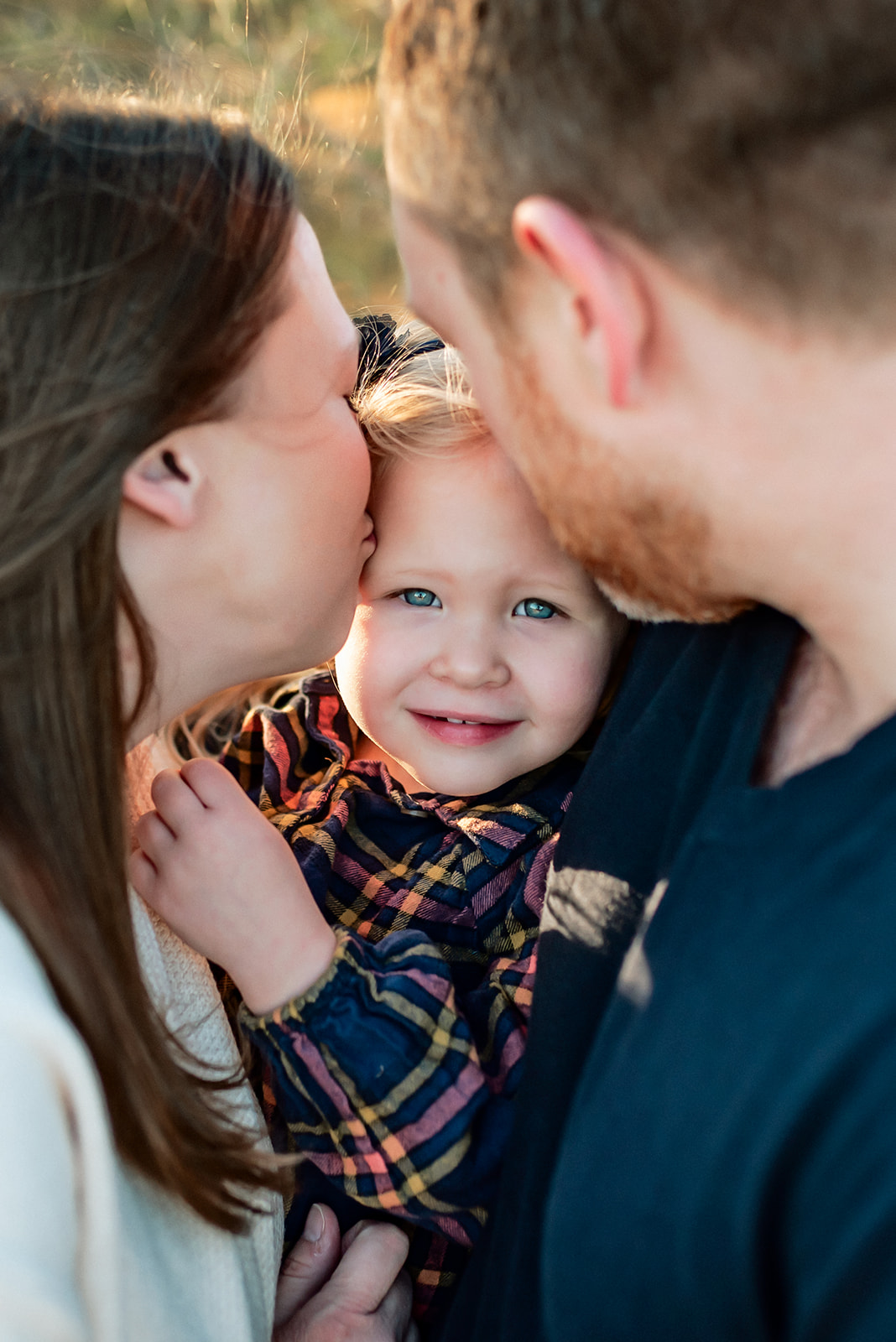 Mom and dad kiss little girl on the head while she's held between them.