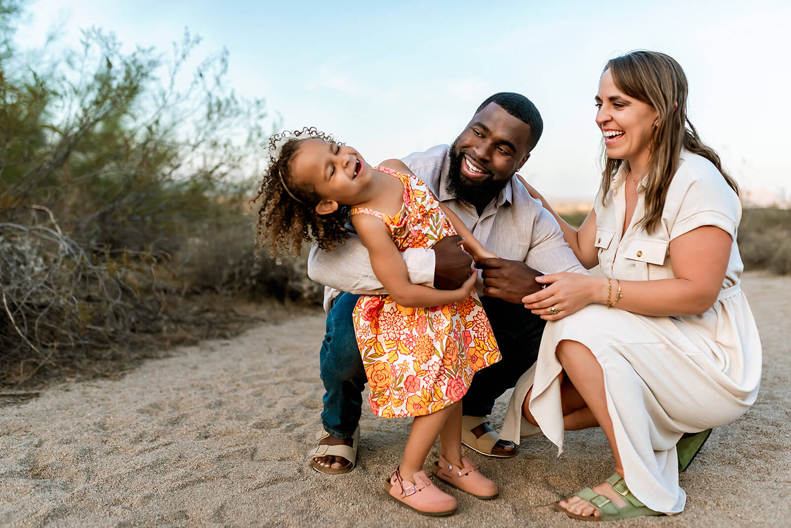 Little girl throws her head back and laughs while her parents kneel down around her during a spring family photo session.