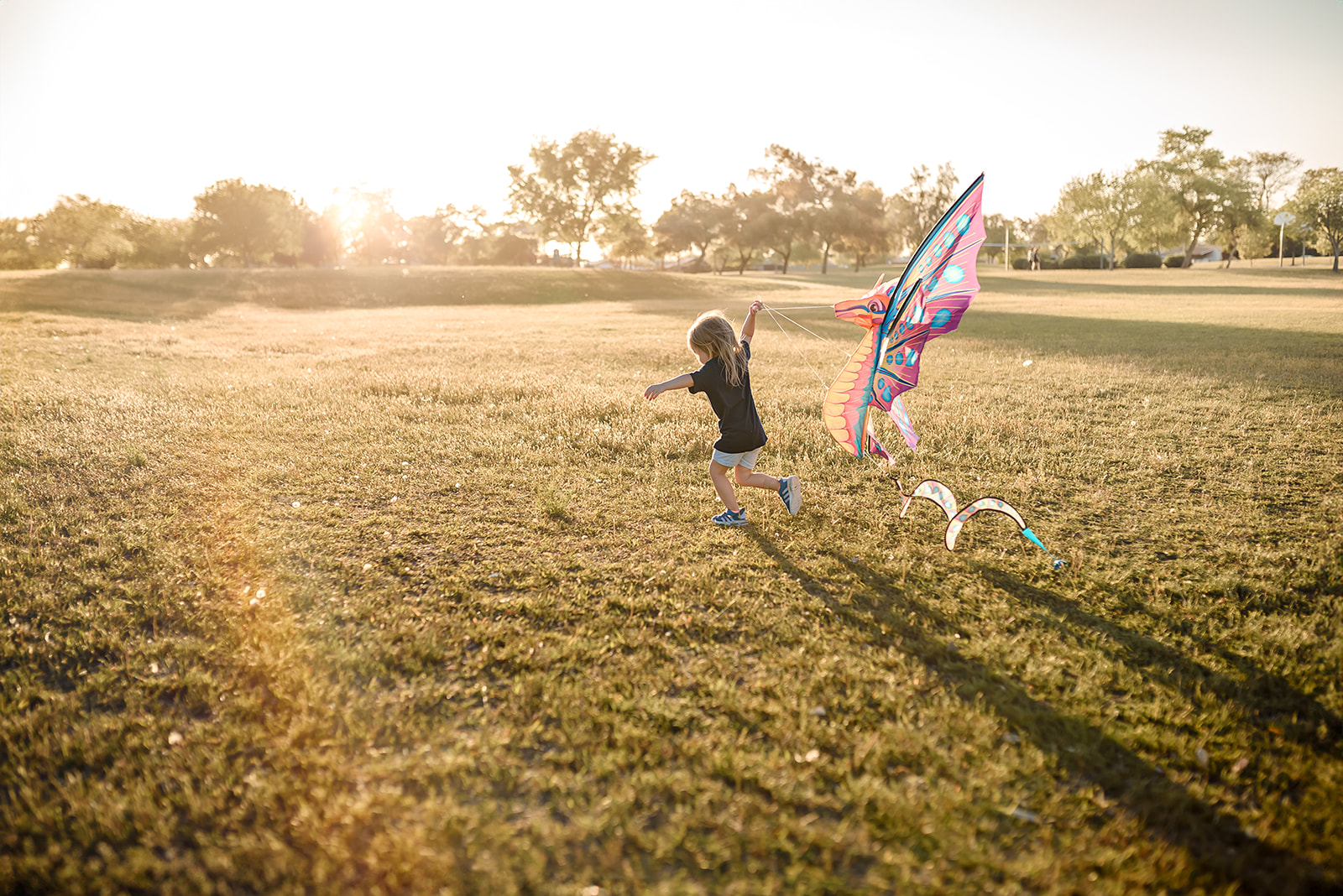 Little girl runs in a field carrying a kite behind her with golden sunset light