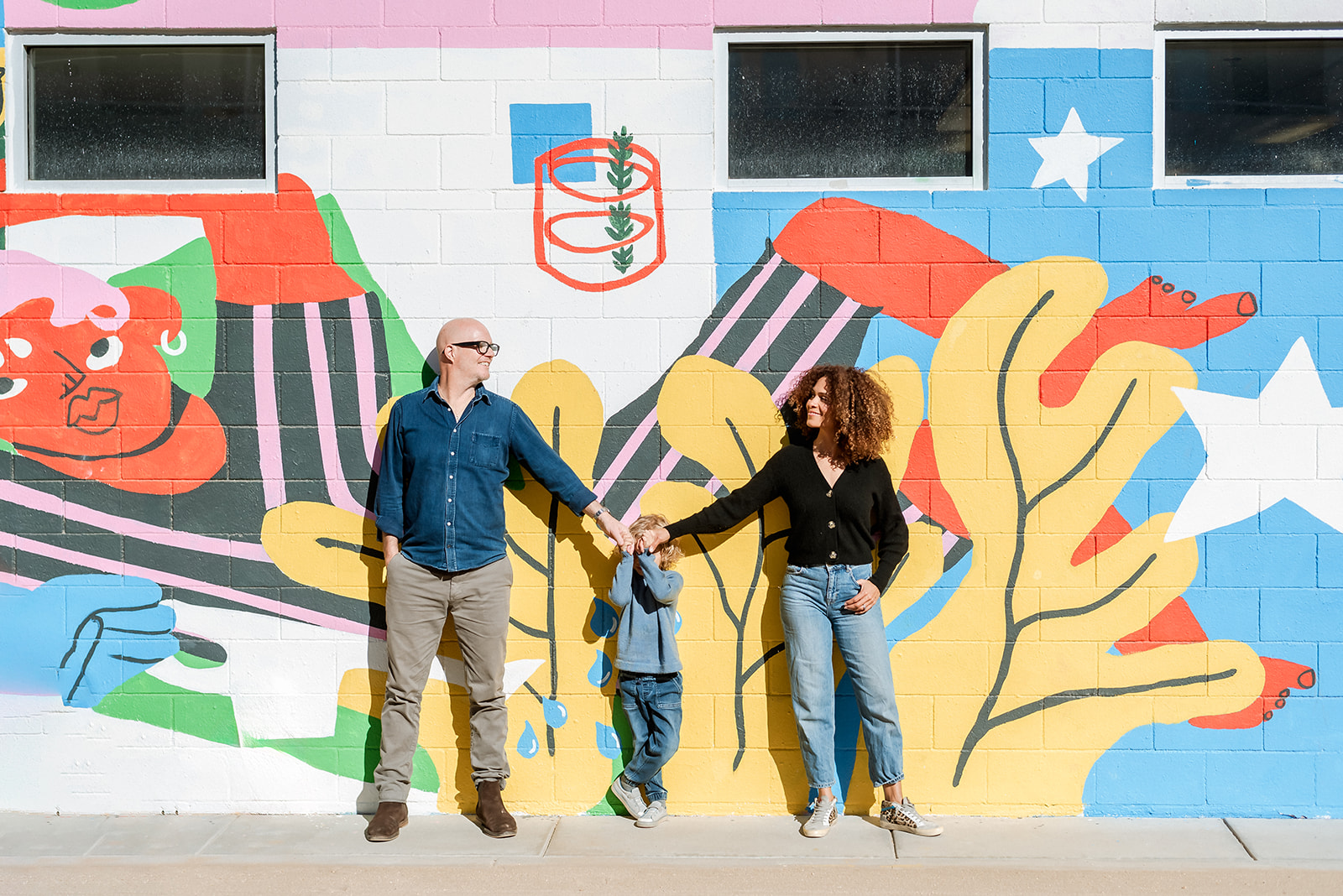 Family of three stand against a brightly painted wall holding hands and looking at each other.