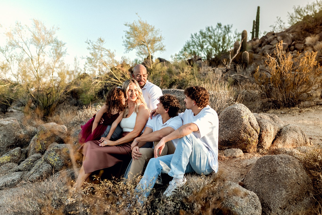 Family of five sits on a group of boulders in the Phoenix desert and smiles at each other.