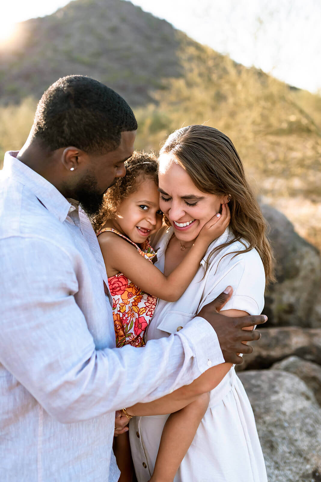 Family of three embrace while mom holds little girl. Little girl places her hand on mom's cheek and dad looks at them lovingly