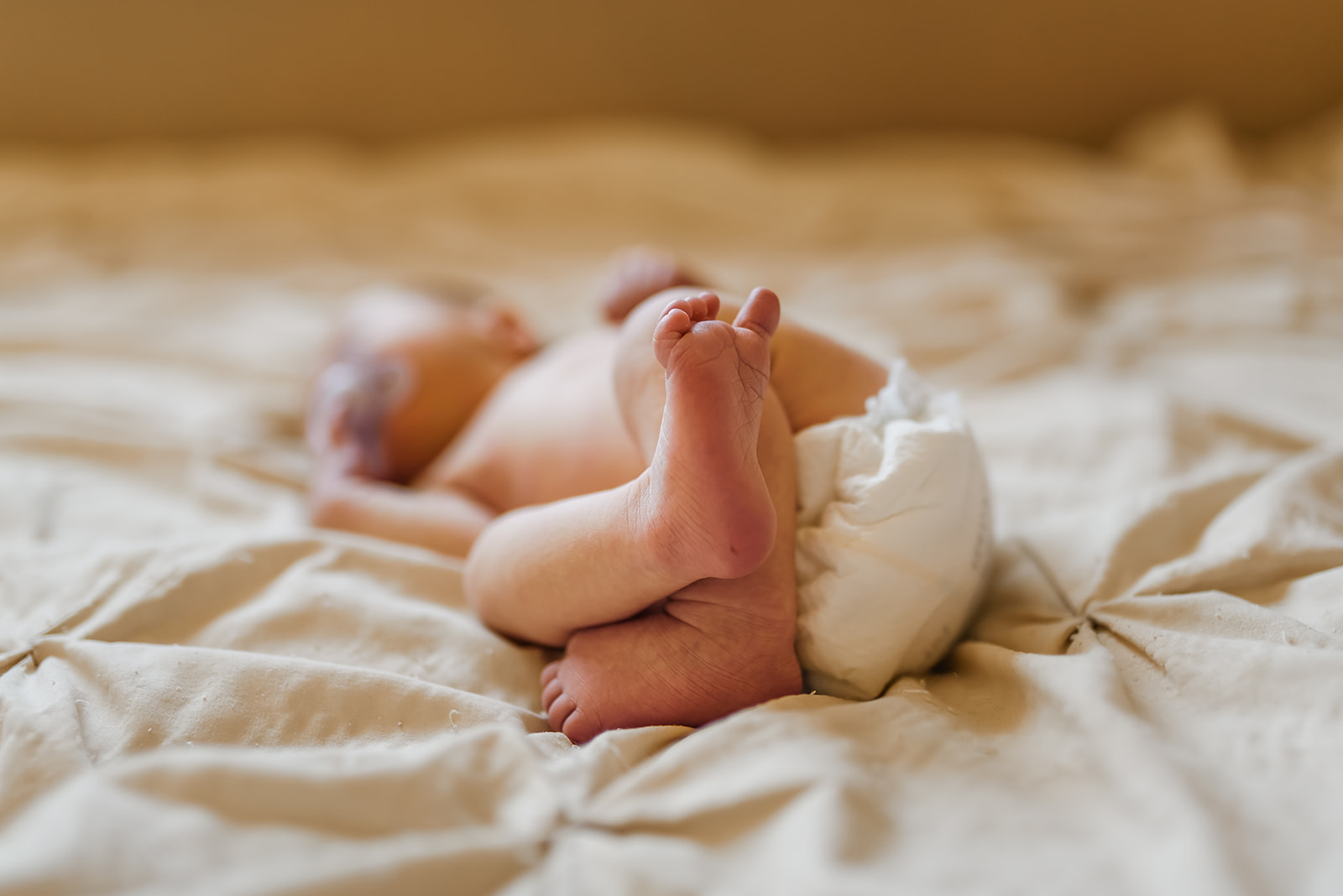 Newborn baby girl laying on a cream colored bed spread with the focus on the baby's feet.
