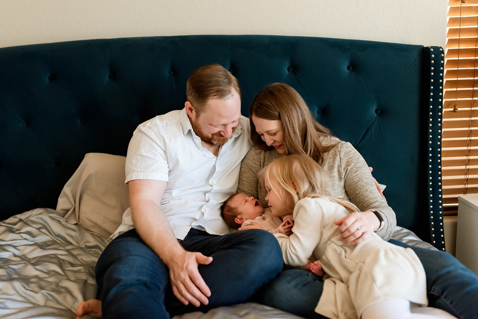 Family of four sit on a bed and snuggle with newborn baby.