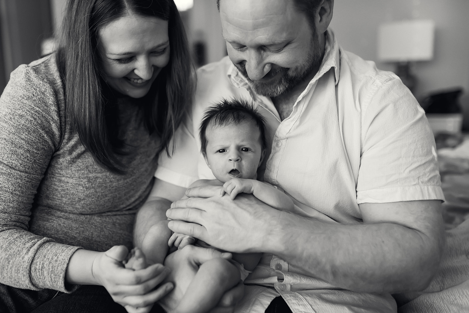 Black and white photo of newborn looking intently towards the camera while dad holds her and moms its next to them.