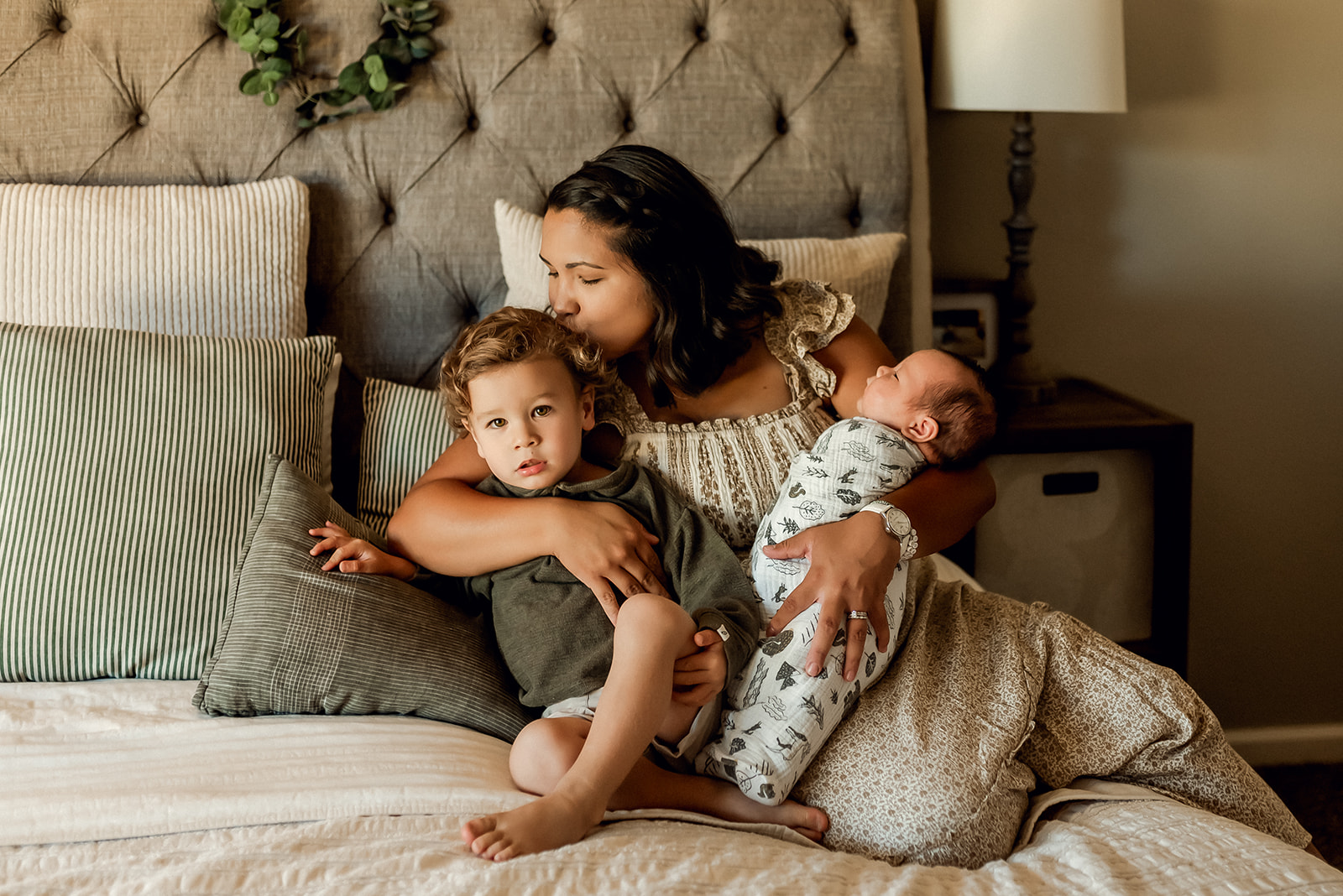 Mom sits on her bed kissing her toddler on the head and holding her newborn baby wrapped in a blanket.
