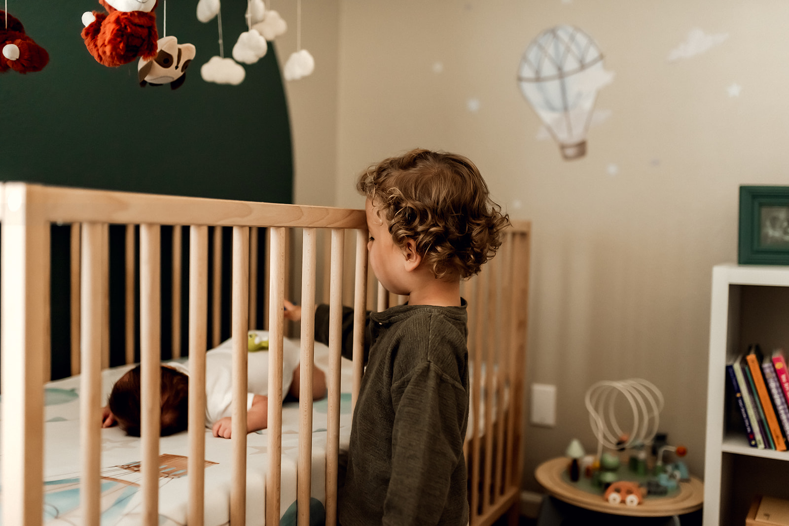 Toddler boy with curly hair stands in front of a crib where his newborn baby brother sleeps.