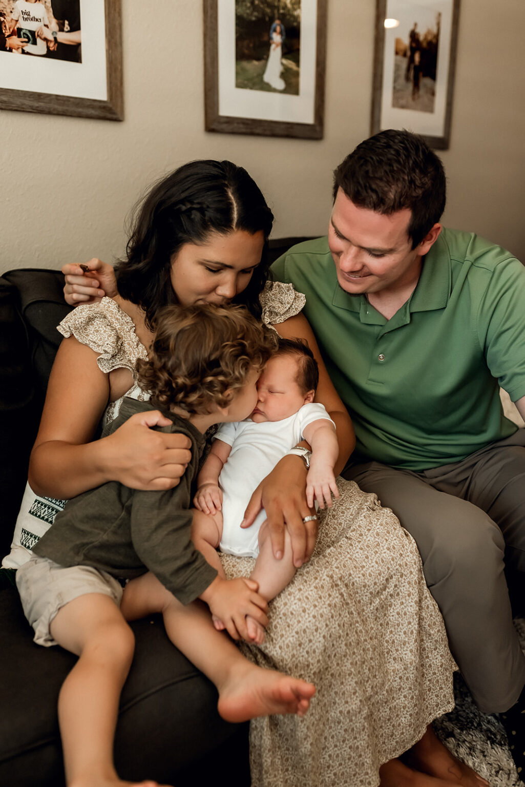 Toddler boy gives newborn baby brother a kiss while mom and dad look at them both lovingly.