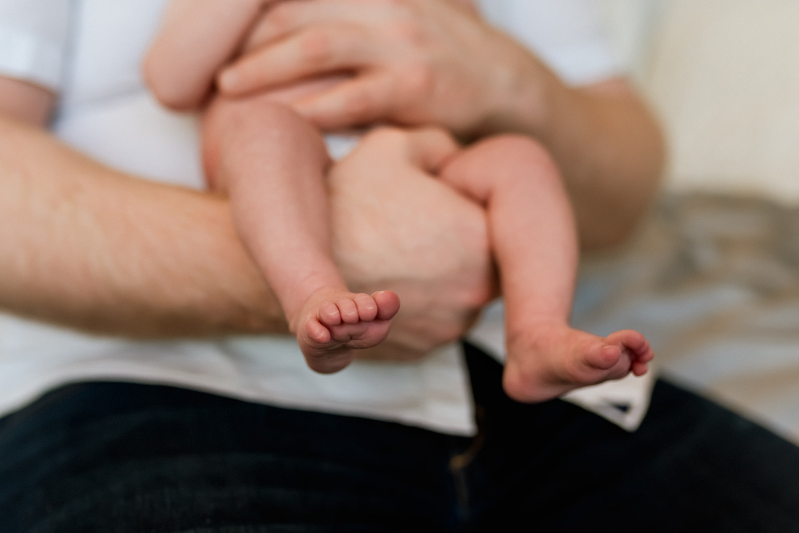 Newborn baby feet and legs dangle while being held their dad's arms.