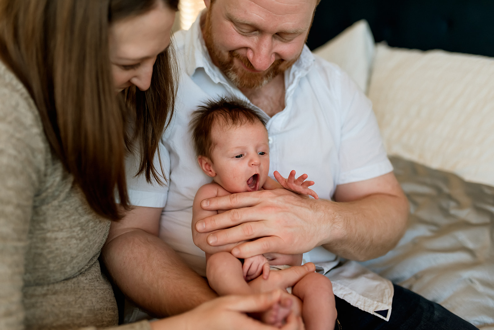 Mom and dad sit together on a bed holding their newborn who yawns