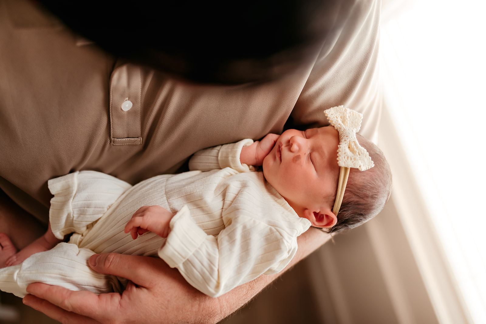 Dad wearing a tan colored shirt holds newborn in one arm while standing near a window.