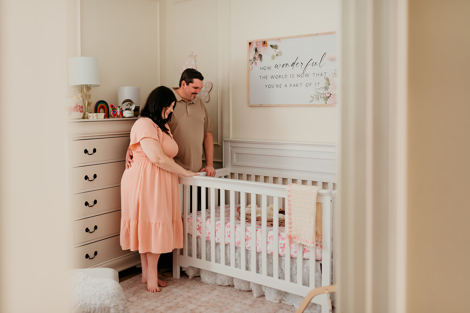 Mom and Dad are framed by a doorway lovingly looking into a crib during an in-home newborn photo session.