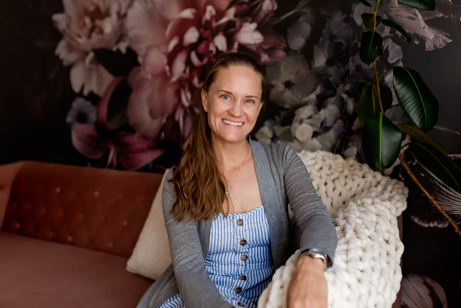 Woman with light brown hair, wearing a striped button up dress and grey sweater smiles at the camera